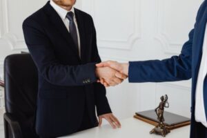 Two professionals in suits shake hands across a desk, symbolizing agreement or partnership, with a small justice statue visible nearby.
