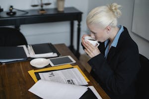 Side view of young concentrated young female judge in formal suit drinking hot coffee while reading case files while sitting at table with stack of documents and tablet